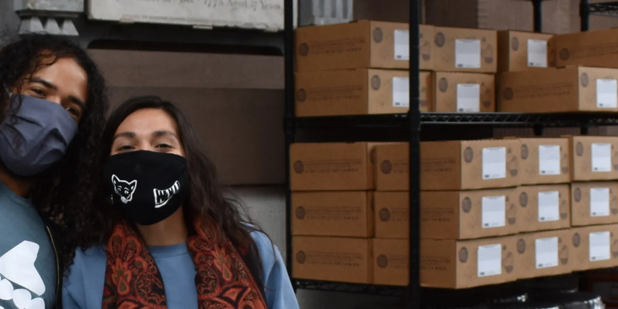 Two people stand in front of a building, the taller one with his hand lifted up in a wave. They stand next to a stack of brown boxes.