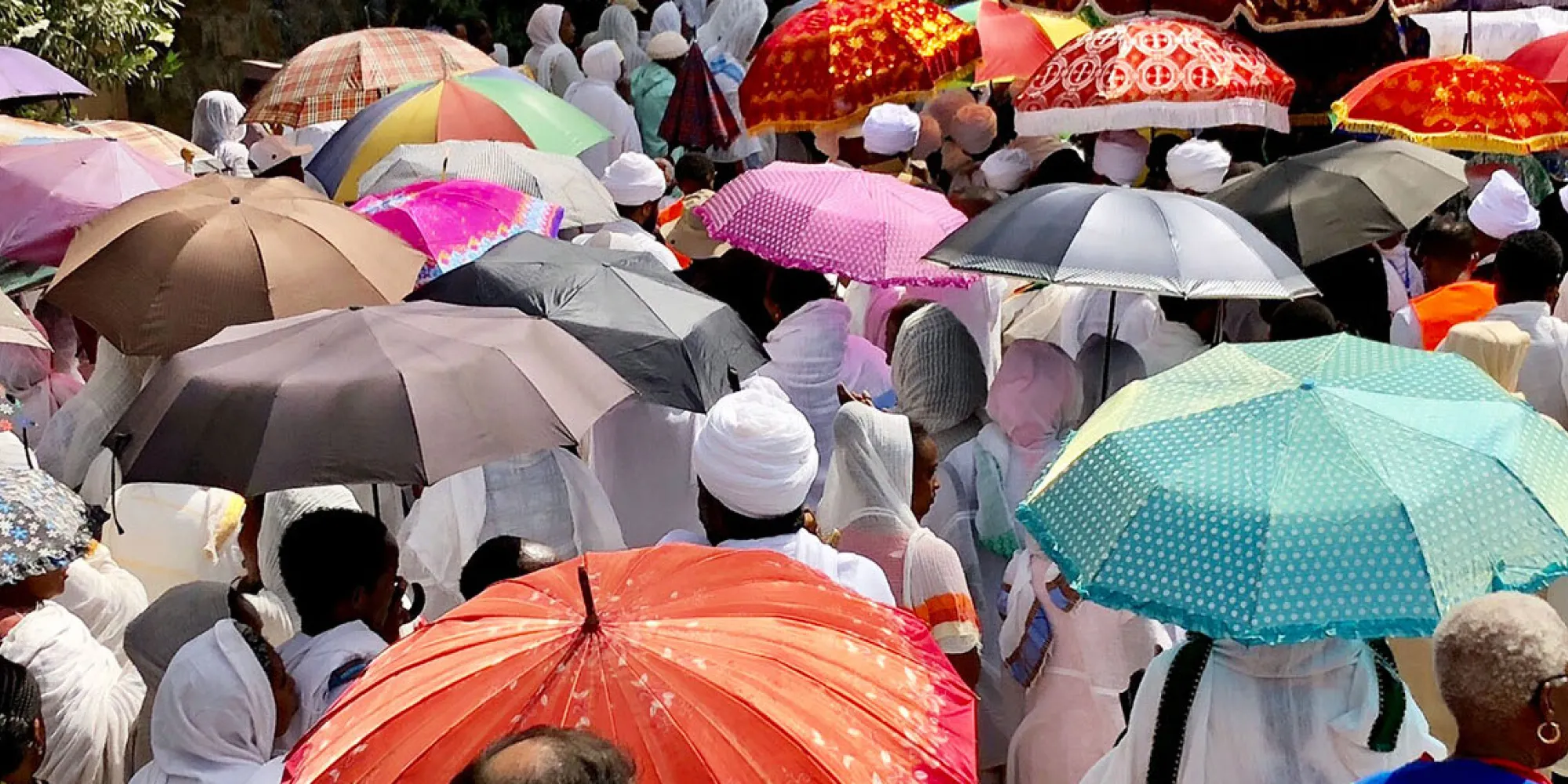 A crowd of people stand on a path. Many hold colorful umbrellas or wear scarves to protect themselves from the sun.