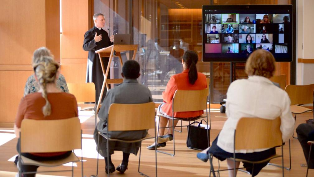 Members of the congregation sit in chairs with WebEx screen and Vicar speaking at Discovery