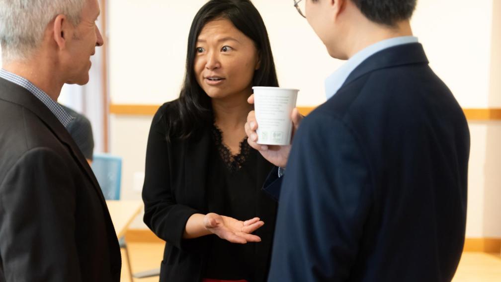 An Asian woman is in conversation with a White man and Asian Man. She has dark hair and is wearing a black cardigan, black lace-lined blouse, and red skirt. The White man has white hair and is wearing a dark grey suit. The Asian man wears glasses, a navy suit, and is holding a white coffee cup.
