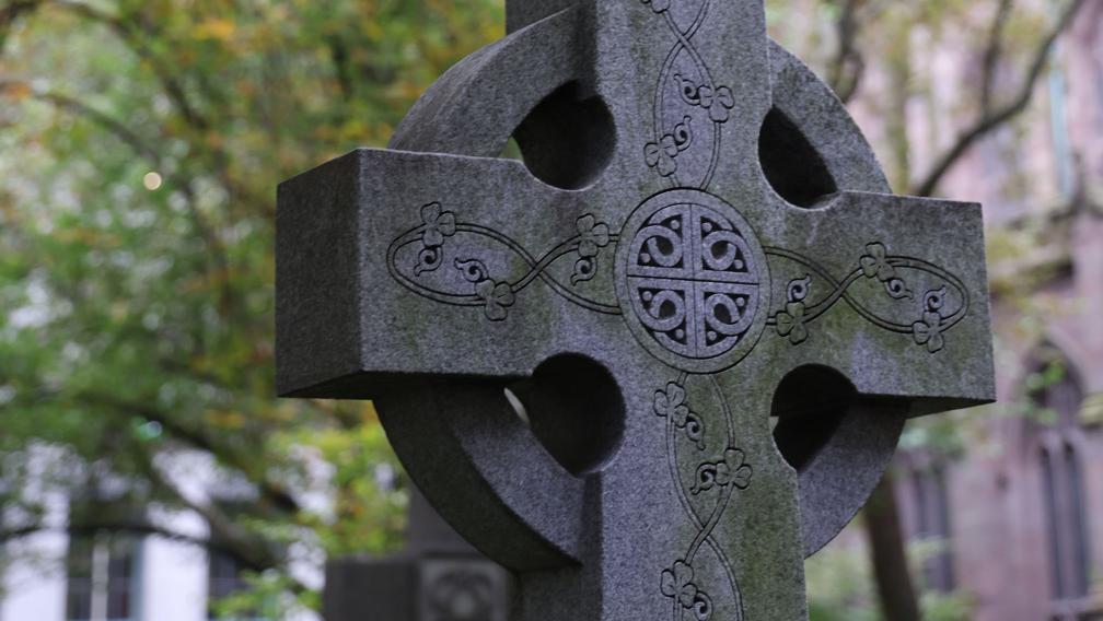 A close-up photograph of a cross-shaped headstone in Trinity Churchyard with trees and Trinity Church in the background