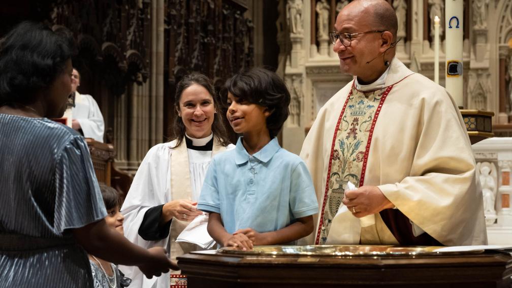 A young baptismal candidate stands at the font with Father Phil and Mother Kristin