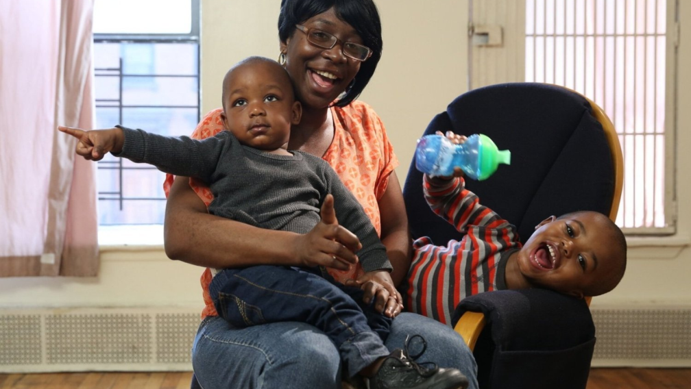 A Black mother wearing glasses and her hair in a black bob smiles with her two children. One child smiles and leans over with his sippy cup in hand, while the other sits in her lap with a serious expression and points to the left.