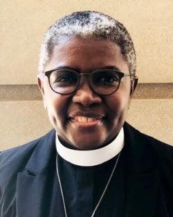 Headshot of The Rev. Sr. Promise Atelon. Sr. Promise wears a white collar, black shirt and jacket, and round glasses. She is smiling, and her dimples are showing in front of a beige wall. Her short, dark hair has some pops of white laced throughout.