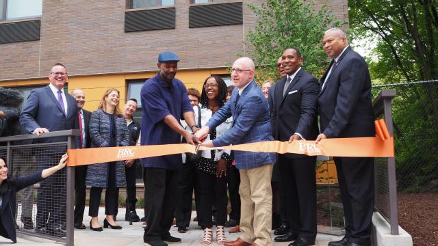 A group of people stand with shears, smiling as they cut a ribbon in front of a building.