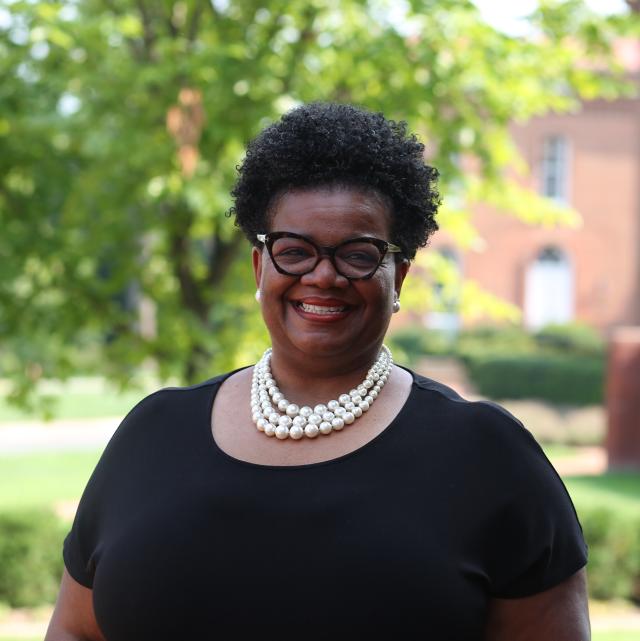 Jacqueline Biallou smiles into the camera. She wears glasses and a three-strand pearl necklace with a black top.