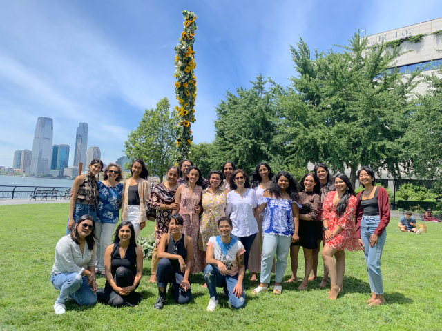 A group of South Asian women stand in a green outdoor space, smiling at the camera. There is a large stalk of sunflowers behind them.