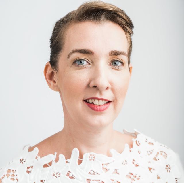 Carey Wallace looks directly into the camera against a light background, wearing a white lace top.