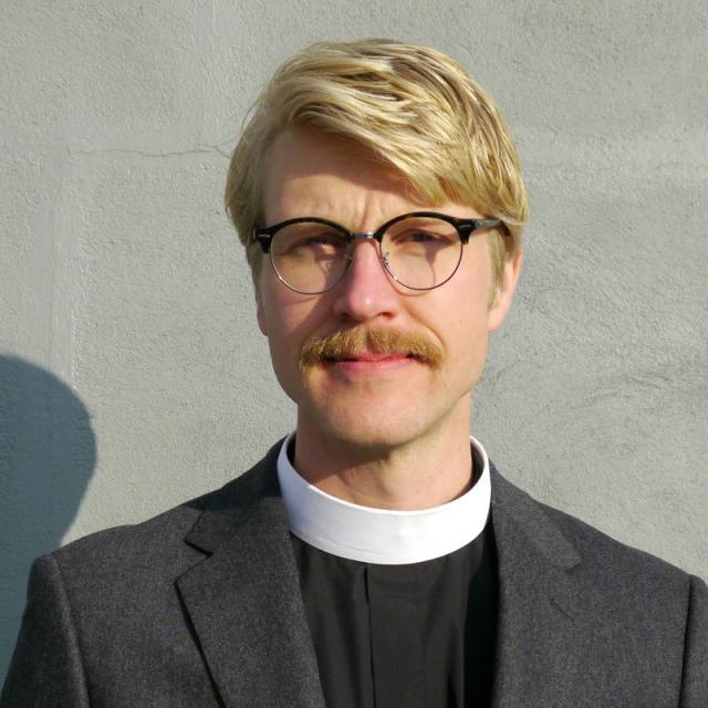 Carl Adair looks directly at camera, wearing clergy attire and a dark grey blazer.