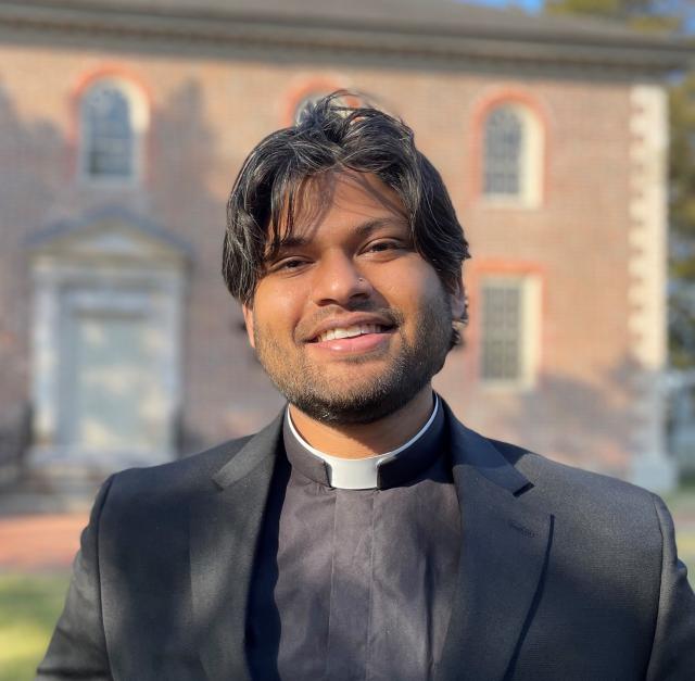 The Rev. Celal Kamran smiles directly at the camera wearing clergy attire, with sunlight shining across his face. In the background, there is a brick building out of focus.