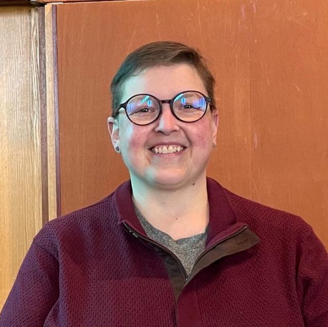 Jen Crompton smiles at the camera while standing in front of a wood-toned wall. They wear a burgundy jacket over a grey shirt and glasses.