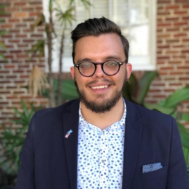 Jordan Smith smiles at the camera, wearing round glasses, a printed button down shirt, and navy blazer with a pocket square and flag pin on his lapel. 