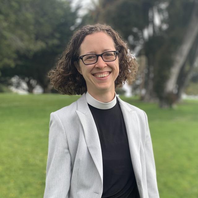 The Rev. Kristin Saylor smiles at the camera as she stand outside, wearing a priest's collar and light grey blazer.