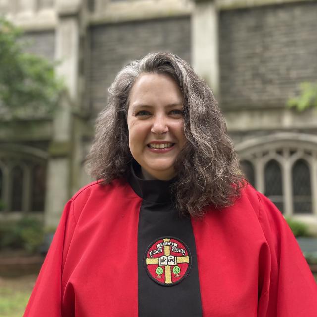 Maryann Pillbrook smiles directly at the camera outside a stone building, wearing a red graduation robe.