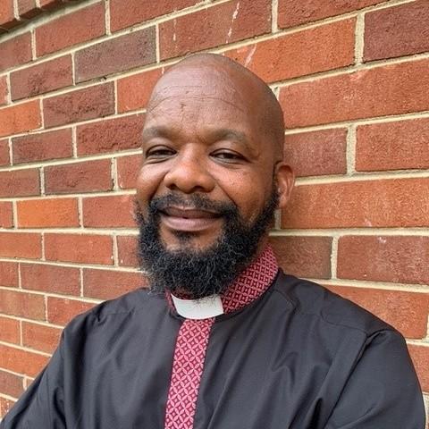 The Rev. Mawethu Ncaca stands in front of a brick wall, smiling softly at the camera. He wears a priest's collar and a shirt with red patterned accents.