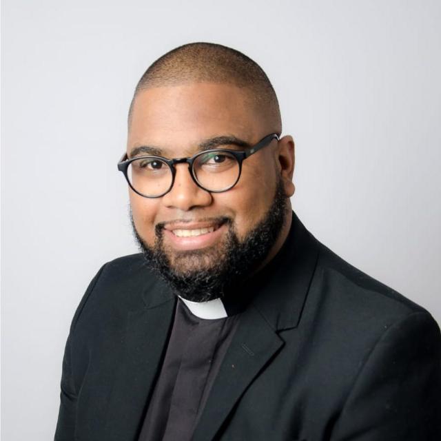 Nitano Muller smiles directly into the camera, wearing clerical garments and a pair of glasses.