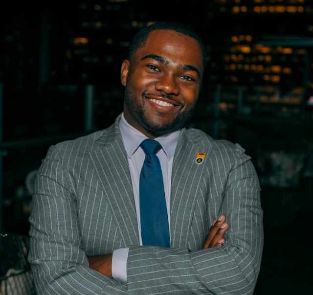 Riley Jones smiles at the camera while crossing his arms and wearing a pinstriped suit.