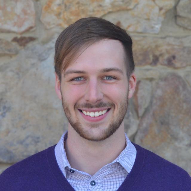 Christopher Schwenk smiles at the camera, wearing a sweater over a button down shirt. He stands in front of a stone wall.