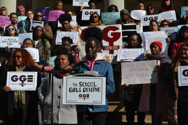 Black girls, teens, and women of varying skintones and hair textures hold signs that read "GGE," "The School Girls Deserve," and "Black girls deserve safe, free, and just schools." 