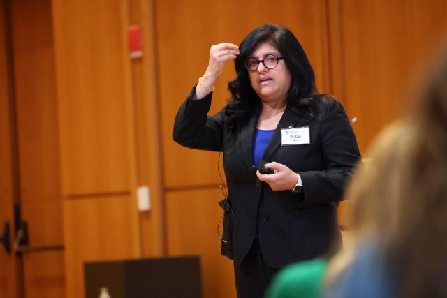 Dr. Gia Merlo speaks with her right hand lifted up. She is wearing glasses with a dark pantsuit and purple blouse. She has dark hair in a bob, with a fair skin.