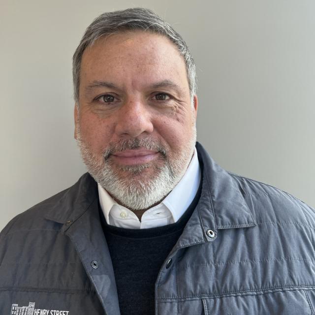 David Garza wears a white oxford, black sweater, and grey quilted jacket. He has grey and white hair, with a white beard and mustache.