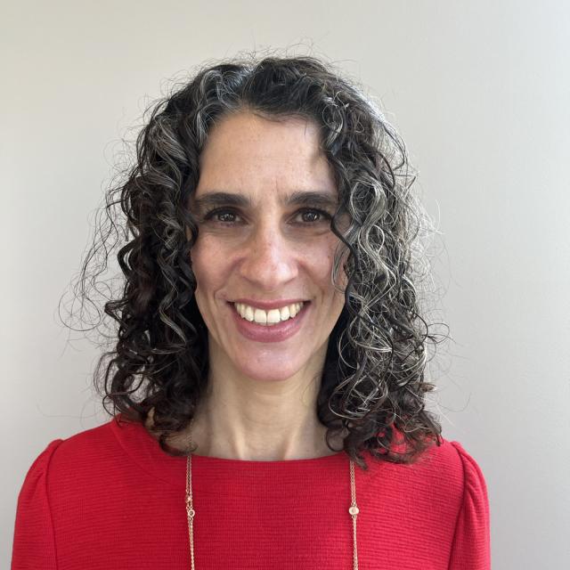 Samara Karasyk smiles, wearing a red top and long gold chain necklace. She is a white woman with salt and pepper curly hair worn down at shoulder length.