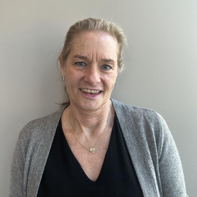 Alison Overseth smiles at the camera. She has blonde hair in a ponytail and wears a black v-neck blouse with a grey cardigan and a delicate gold necklace.