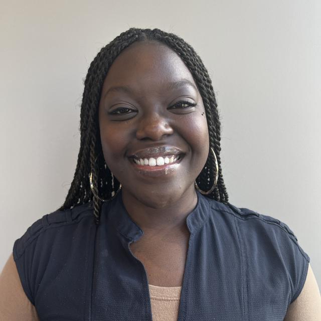 Ebonie Simpson smiles at the camera, wearing a khaki long-sleeved tee under a navy blouse. She is a dark-skinned Black woman with dark brown braids, wearing silver hoop earrings.