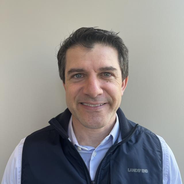 Darren Bloch smiles at the camera wearing a light blue button down and navy Land's End vest. He has dark brown hair cut short with a widow's peak, and light blue eyes.