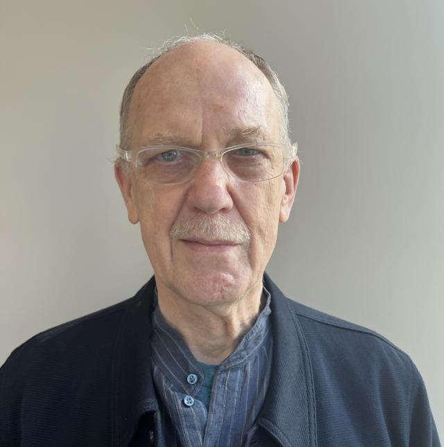 David McCorkle wears clear-framed glasses, a striped blue collarless shirt over a blue undershirt, topped with a navy collared jacket. He is a Caucasian man with light blue eyes, balding white hair, and a mustache.