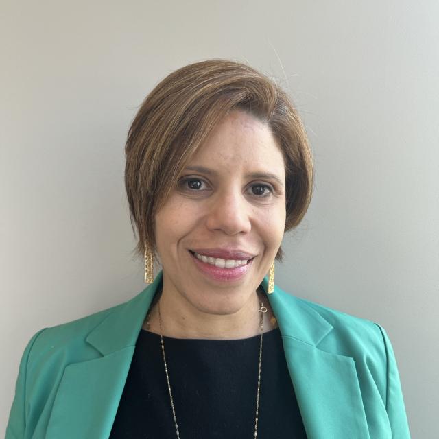 Gisele Castro smiles at the camera, wearing a kelly green blazer over a black boatneck blouse. Her highlighted hair is cut in a bob and she wears rectangular gold earrings with cut outs and a long gold necklace.