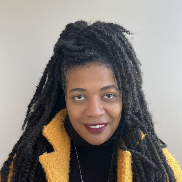 Jenna Chrisphone smiles at the camera, wearing a black turtleneck, yellow coat, and burgundy lipstick. She is a Black woman with medium brown skin and hair in twists, worn half up and half down.