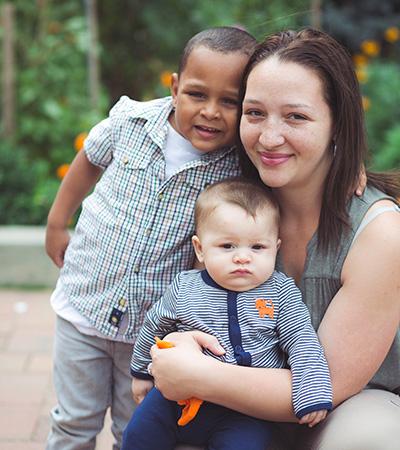 A young Latina woman with freckles and dark shoulder-length hair embraces her two small children and softly smiles at the camera.