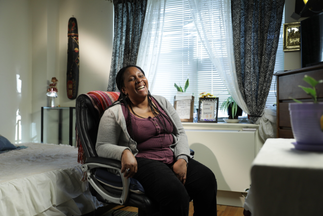 A Black woman with braids laughs while seated in a desk chair in her home. She wears a grey cardigan, purple blouse, and black pants. 