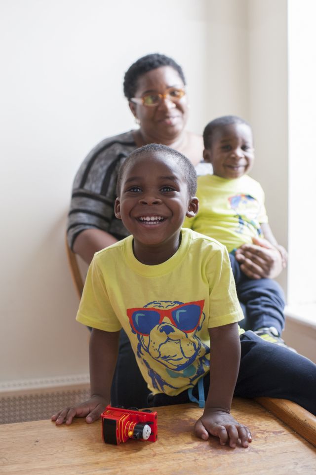 A Black mother wearing glasses and a striped shirt holds her younger child in her arms in the background. In the foreground, her older son smiles wide at the camera, wearing a yellow shirt with a decal of a bulldog wearing sunglasses on it. A red Thomas the Tank engine toy lays sideways in front of him.