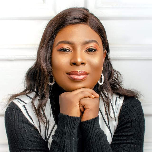 Adekemi Adeniyan smiles with her hands clasped under her chin. She wears a black ribbed long-sleeved top, with a white and black striped vest and white hoop earrings.