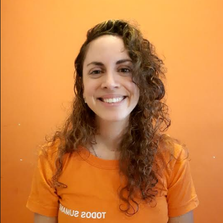 Ana Argento Nasser smiles while standing in front of an orange backdrop wearing an orange shirt. Her hair is curly.