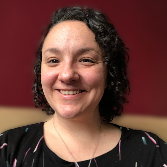 Brenna Davis sits in front of a deep red wall, smiling at the camera wearing a black blouse with abstract lines on it and silver necklace. She is a white woman with dark chin-length curly hair and dark eyes.