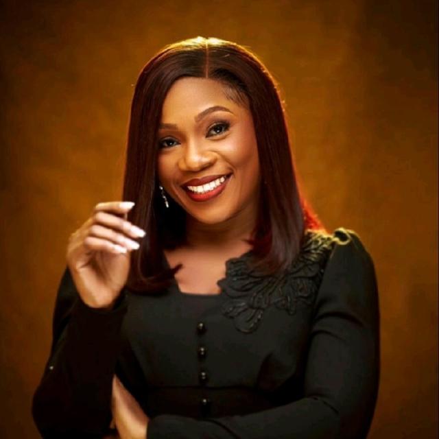 Folake Oni stands in front a warm amber background, smiling brightly at the camera with her hand lifted with fingers slightly curled in front of her shoulder. She is a black woman with a warm brown complexion. She wears her hair in a long bob with red lipstick and a dangly earrings, with a black blouse with slightly puffed sleeves.