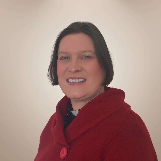 Jennifer Holden smiles at the camera standing a quarter right to the camera in front of a beige backdrop. She is a white woman with dark hair cut into a short bob, with a ruddy complexion. She wears a red sweater with a shawl collar over her priestly collared shirt.