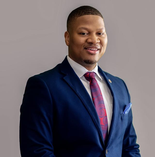 Kenneth Padget smiles at the camera in front of a grey backdrop. He is a black man with short-cropped hair and medium brown complexion. He wears a deep blue suit, white collared shirt, and a red and blue oversized paisley tie.