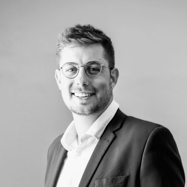 Louis van der Riet smiles directly at the camera in a black and white headshot. He is wearing round glasses, a white collared shirt, and dark blazer. He is a white man with sandy blonde hair.