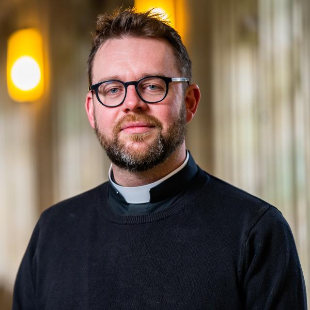 Michael Robinson softly smiles at the camera, with St. Edmundsbury's Cathedral out of focus in the background. He is a white man with a ruddy complexion and dark brown hair and a slightly greying beard and mustache. He wears rounded dark glasses and a black priest's shirt with collar.