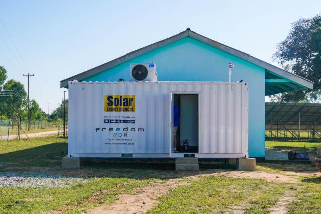 An external shot of machinery in support of the solar farm, featuring a small light blue structure, with a white-painted trailer that says "Solar Energy Projects" written on it. The door to the trailer is open and a male person's back can be seen; he wears a blue top, tan shorts, white socks, and black shoes.