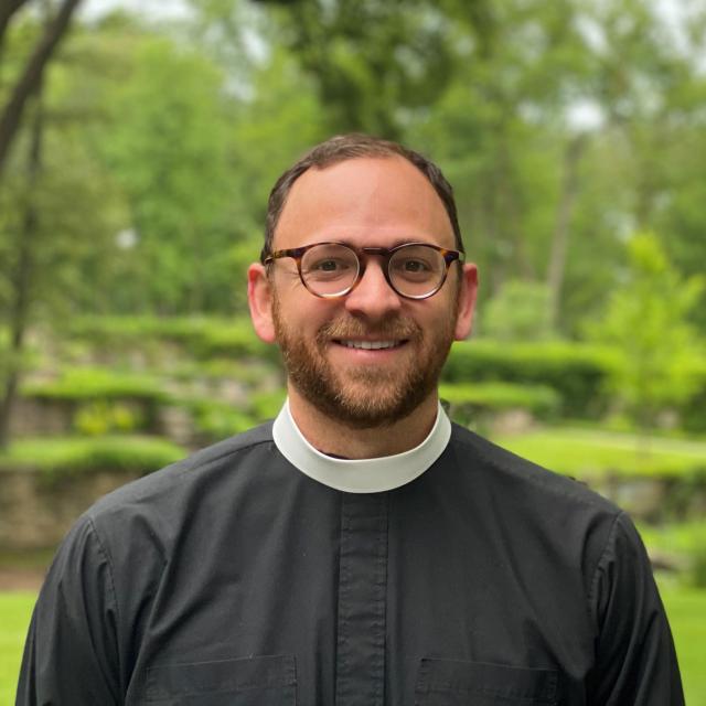 The Rev. Matthew Babcock smiles in the foreground while the background is a verdant outdoor landscape out of focus. He wears a clergy collar and glasses.