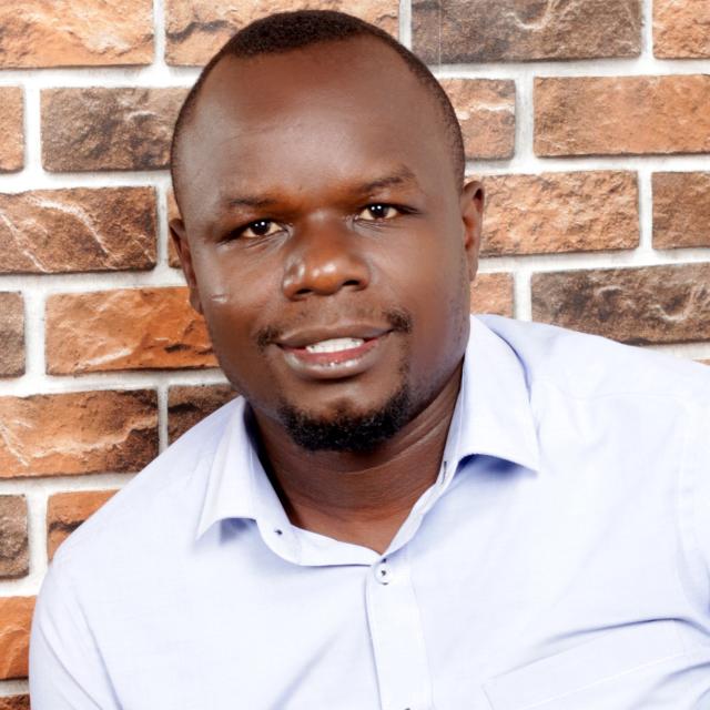 Nicholas Pande smiles as he stands in front of a brick backdrop. He is a black man with short cropped hair and a goatee, with a medium dark complexion. He wears a light blue button up with an open collar.