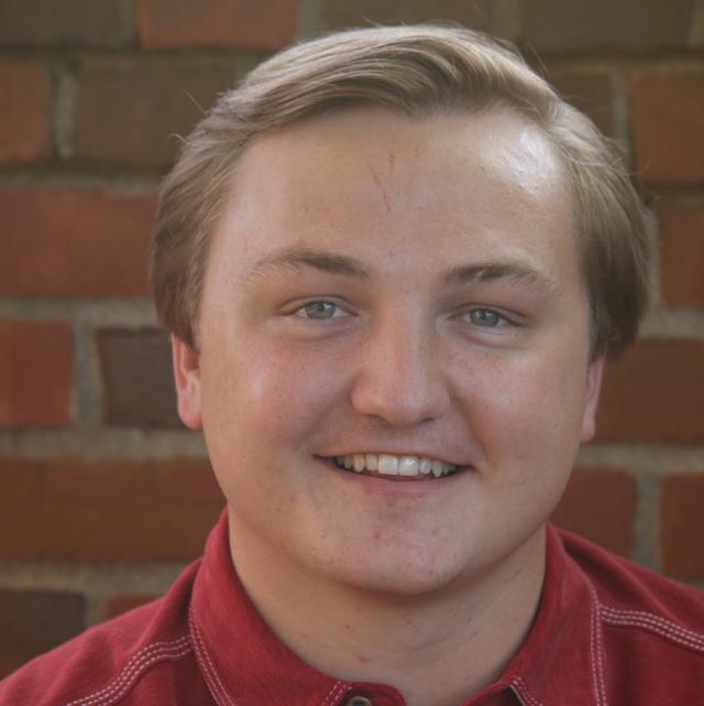 Trey Phillips smiles while standing in front of a brick wall. He is a white man with blonde hair, blue eyes, and a golden complexion. He is wearing a red collared shirt.