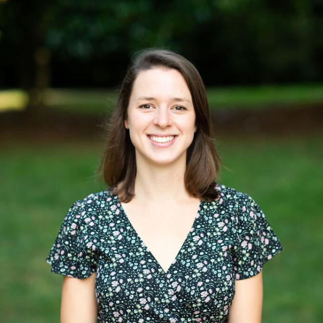 Peyton Williams smiles broadly at the camera, with the verdant grounds of St. Paul's Episcopal Church in the background. She is a white woman with dark brown shoulder length straight hair and creamy complexion and dark brown eyes. She wears a dark blue ditsy floral print dress with a v-neckline.