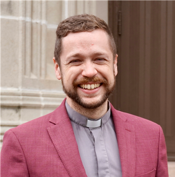 Paul Richards-Kuan smiles while standing in front of a stone-clad church. He is a white man with a pinkish complexion and sandy brown hair. He wears a maroon blazer over grey shirt with a priest's collar.