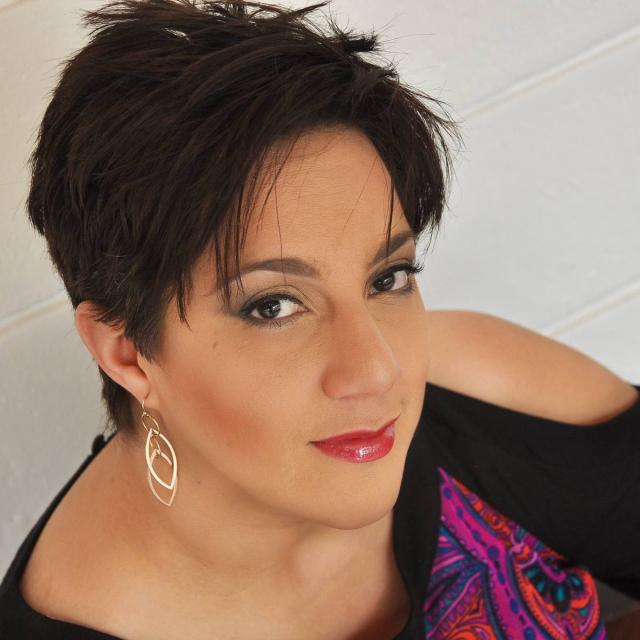 Yuri Rodríguez-Laureani softly smiles up at the camera in front of a white textured background. She is of Latine descent with a golden complexion, short dark hair, and dark brown eyes. She wears make up, gold dangly earrings, and an open-shoulder black blouse with a colorful paisley print at the front.